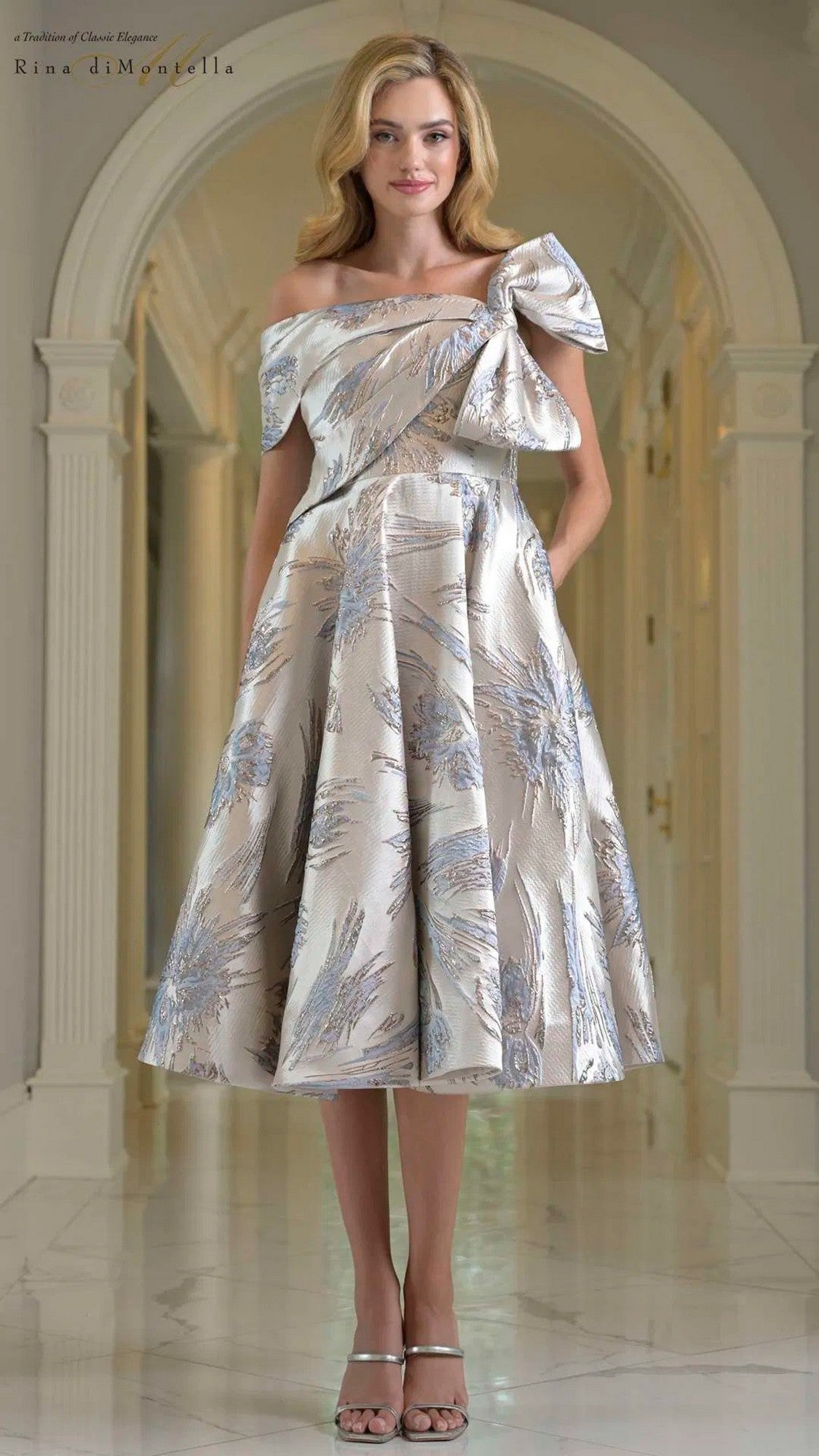A woman stands in a marble hallway wearing the Rina Di Montella RD207, a knee-length semi-formal dress in blue multi with off-the-shoulder style, large bow detail, and elegant jacquard pattern.